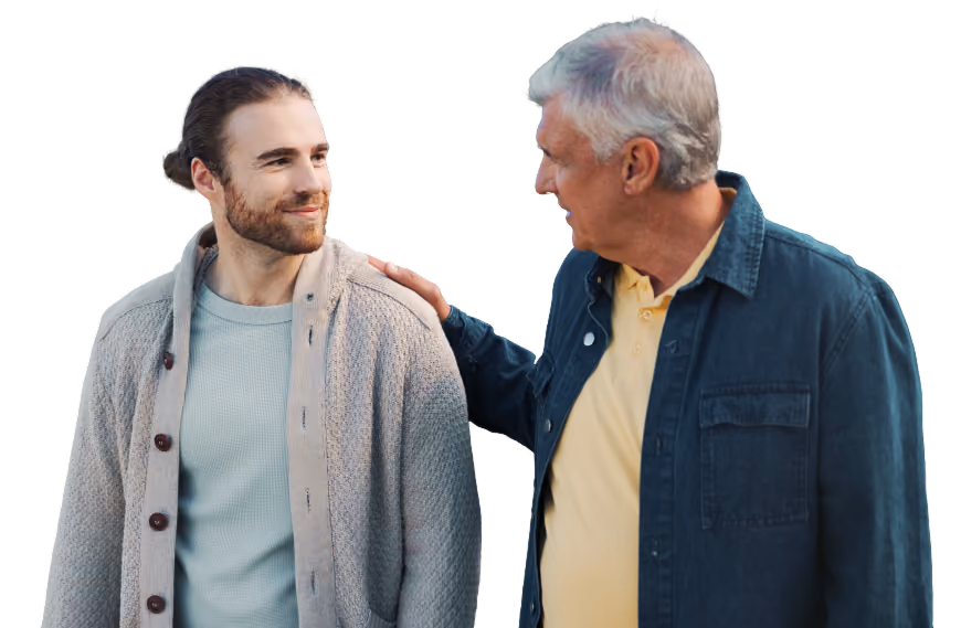 Two male adults standing outdoors in front of a green, tree-filled background. One adult rests a hand on the other adult's shoulder while they face each other, wearing casual layered clothing.