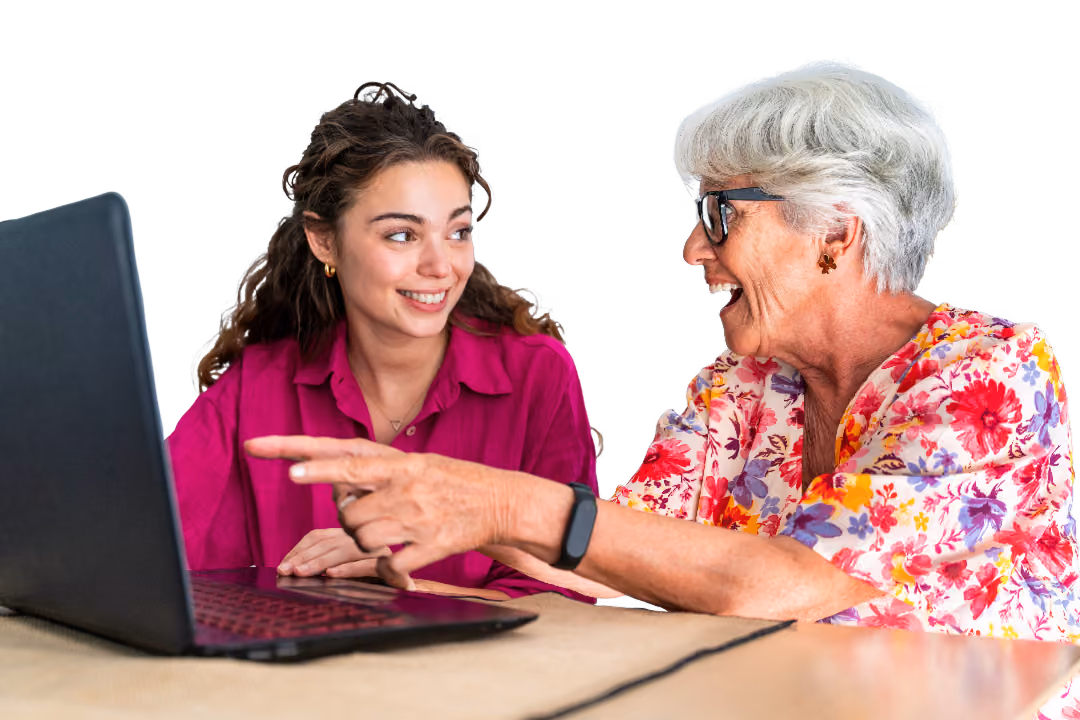 Older adult with short grey hair and glasses sitting beside a younger adult at a table, both looking at a laptop screen. The older adult is pointing towards the screen and wearing a floral shirt, while the younger adult is wearing a bright pink blouse.