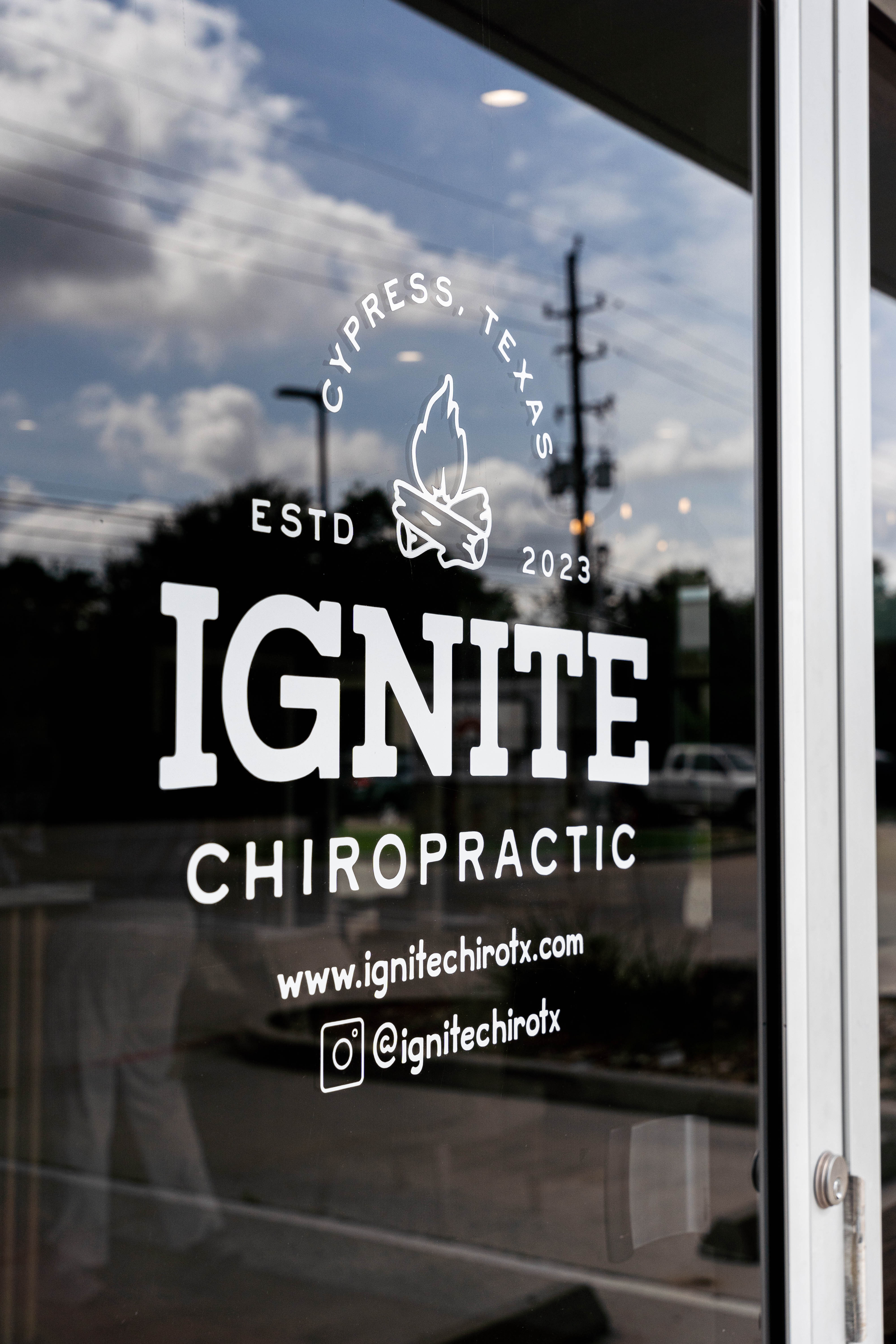 Glass door of Cypress chiropractic clinic with logo, contact info, reflecting sky and street, promoting local healthcare a...
