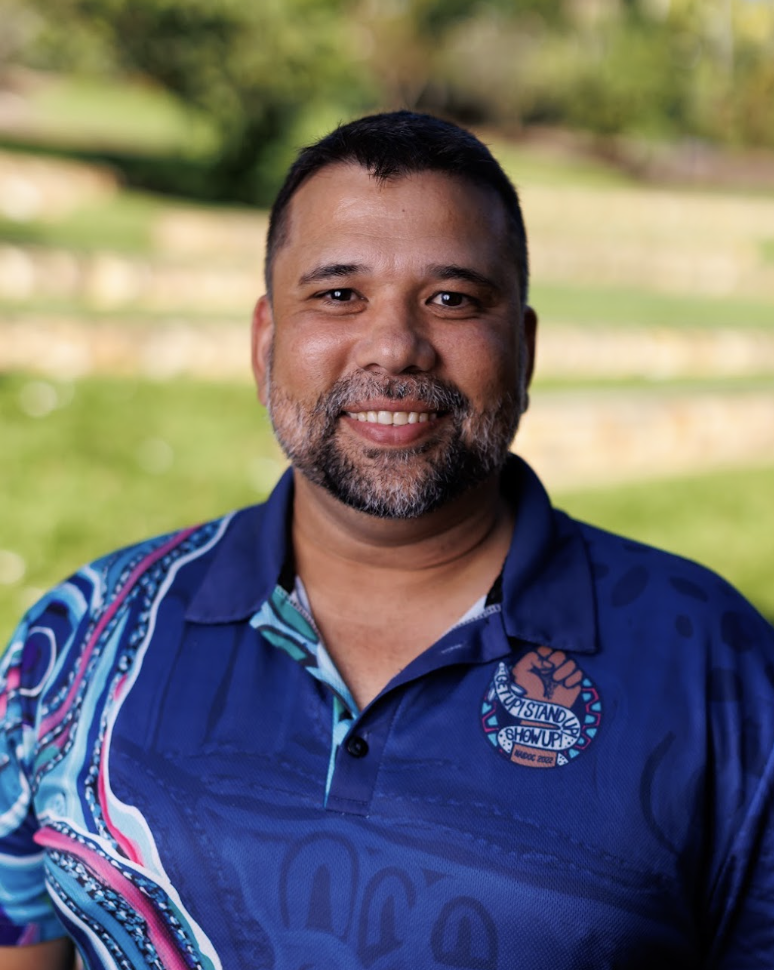 Headshot of Troy Garling wearing a blue decorated shirt with blurred grass in the background.