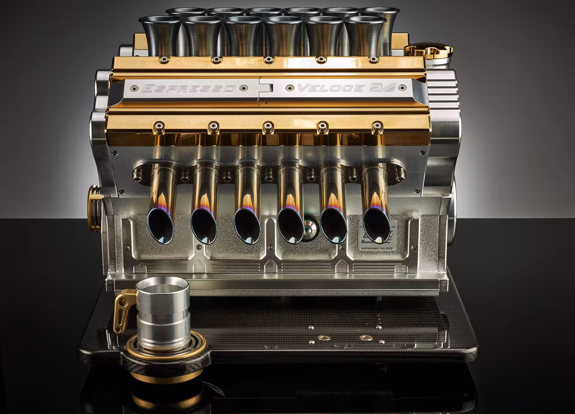 Metal espresso machine front with seven polished spouts and gold accents on black reflective surface.