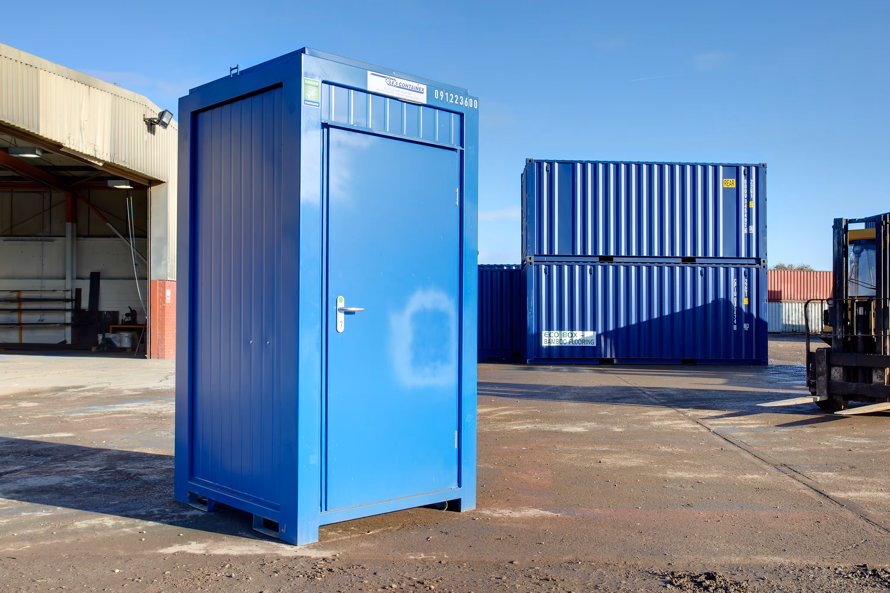 A blue portable toilet is sitting in front of a blue shipping container.