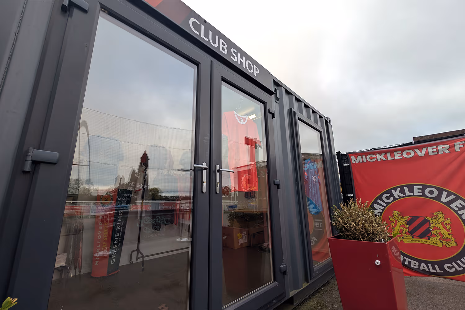 Exterior view of a black portable club shop with sports jerseys visible through glass doors and a red Mickleover Football Club banner nearby.