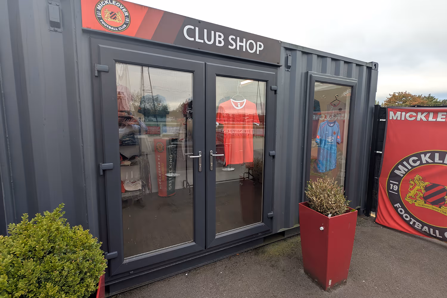 Exterior view of Mickleover Football Club shop in a grey container with glass doors and windows displaying football shirts.