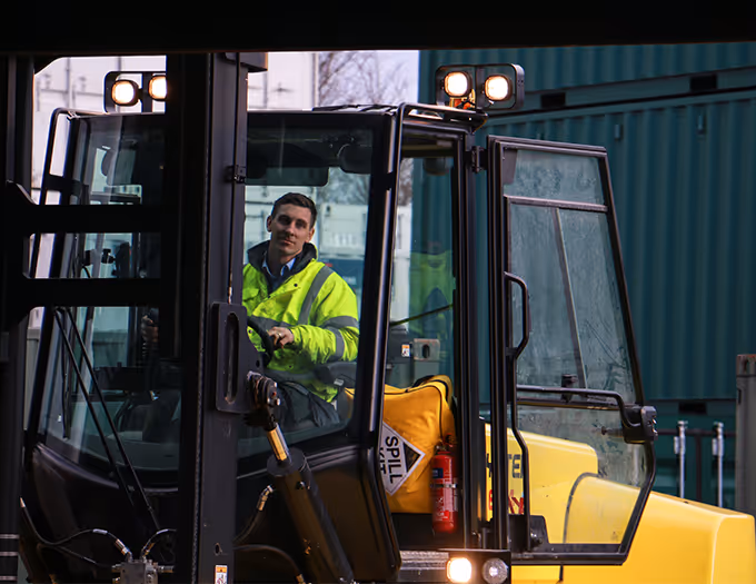 Man in a high-visibility jacket operating a yellow forklift at a shipping container yard.
