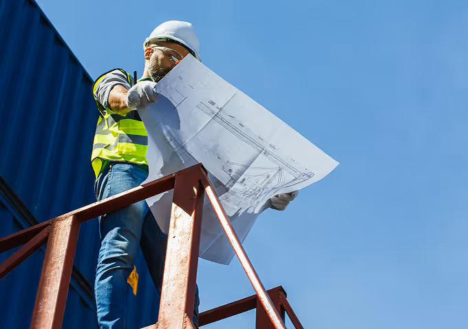 Construction worker in a safety helmet and vest reviewing blueprints outdoors against a clear blue sky.