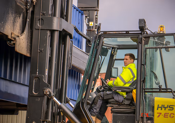 Worker in a high-visibility jacket operating a Hyster forklift lifting a blue shipping container.