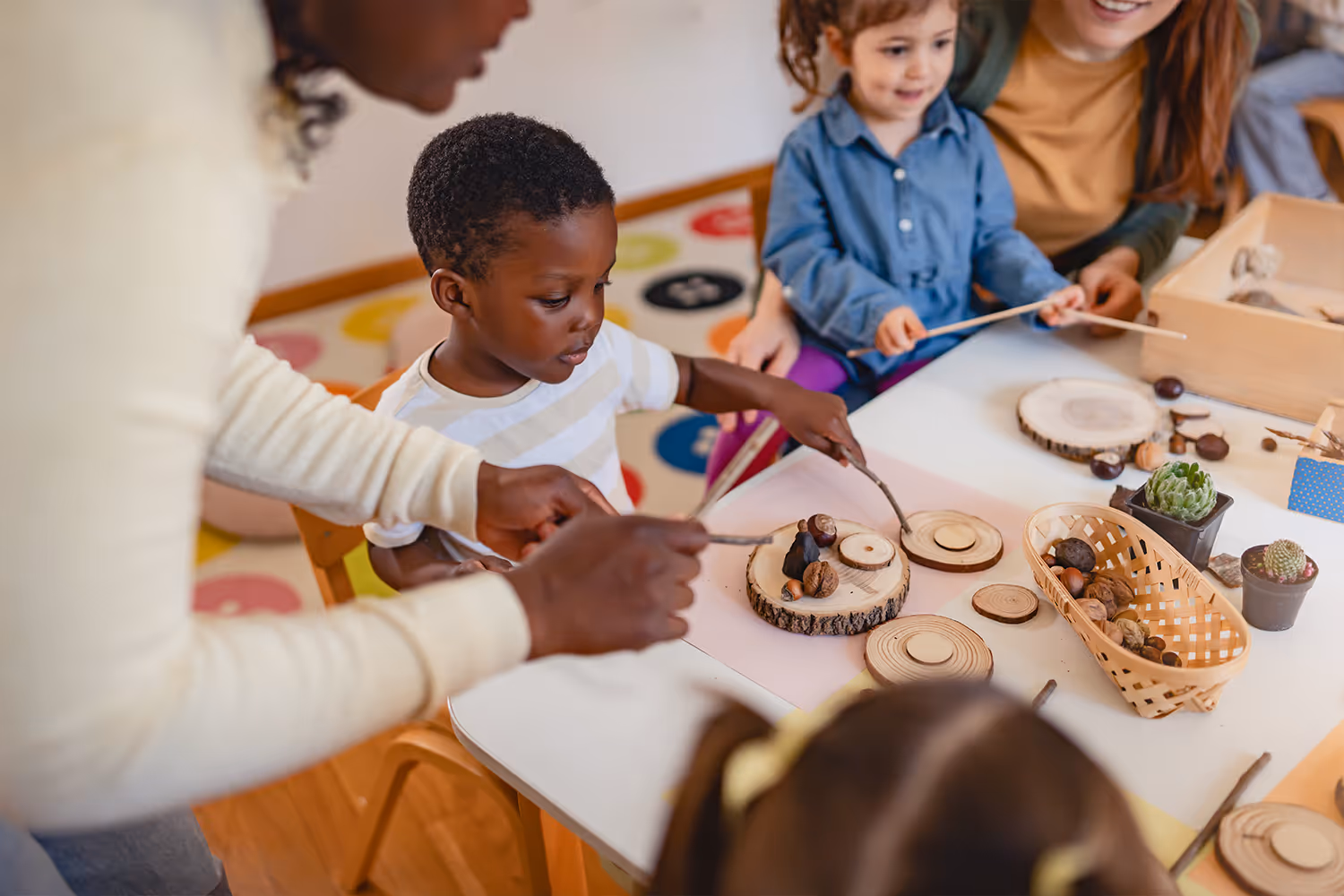 A teacher and young children engaging in a sensory activity with wooden discs, sticks, nuts, and small plants at a classroom table.