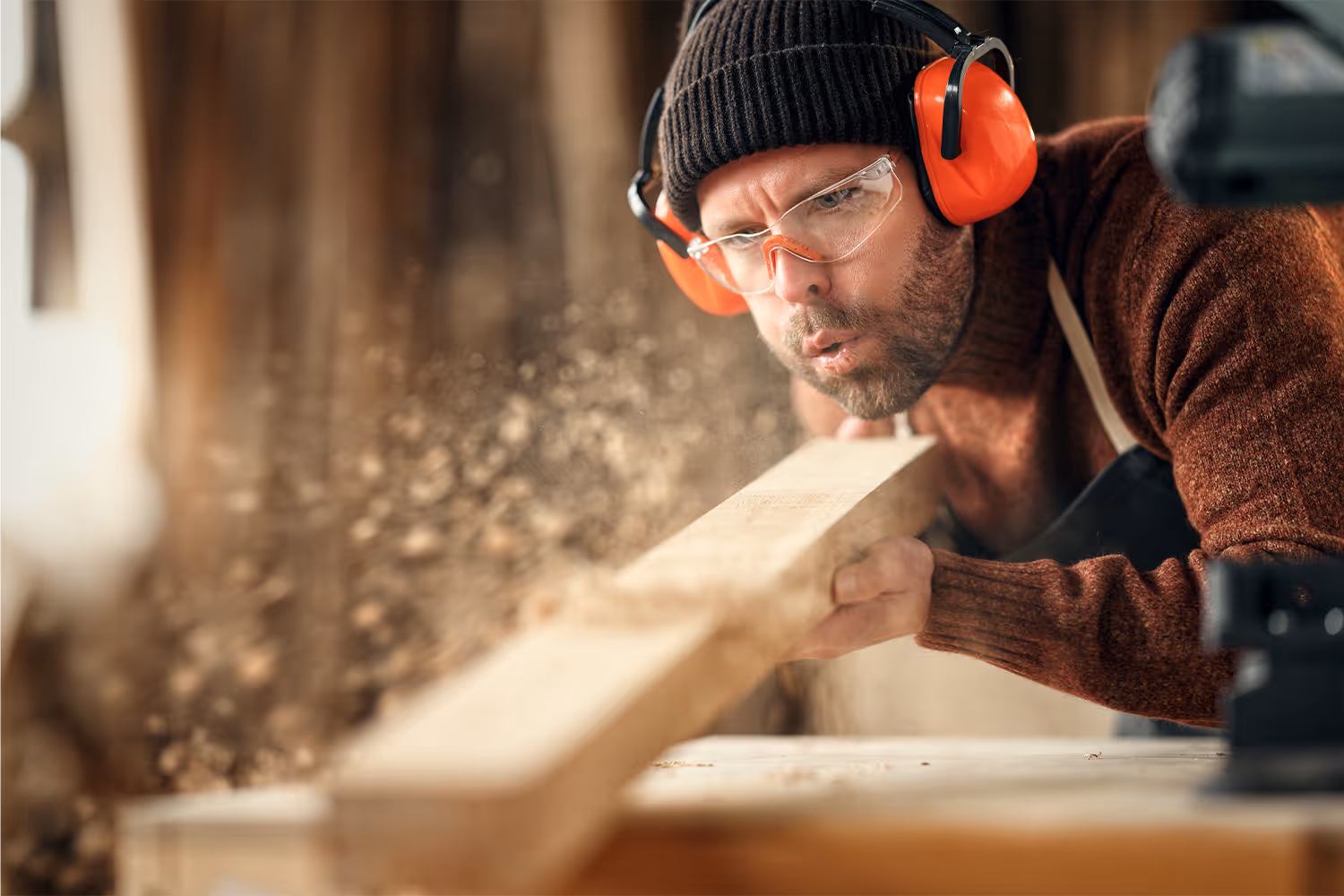 Carpenter wearing safety glasses and ear protection blowing sawdust off a wooden plank in a workshop.