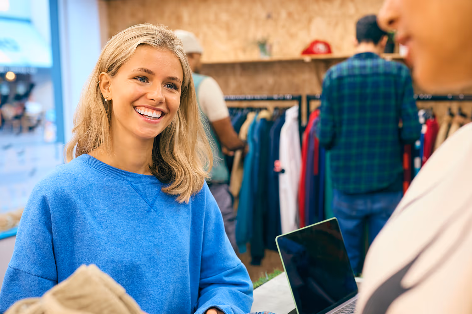 Smiling young woman in a blue sweater holding folded clothes at a retail store counter with a laptop nearby.
