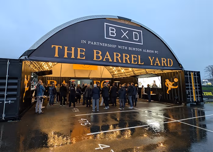 Group of people gathered inside a large black and yellow event tent labelled 'BXD In Partnership with Burton Albion FC The Barrel Yard' on a wet evening.