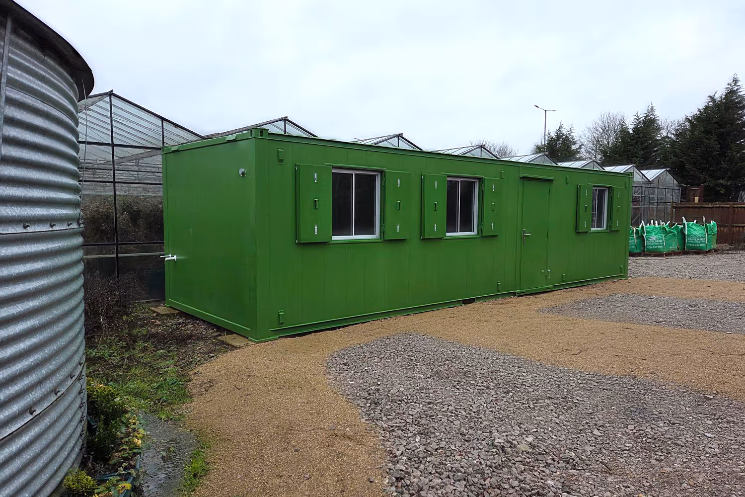 Green portable building with shuttered windows and a door, located beside a corrugated metal structure and greenhouses.