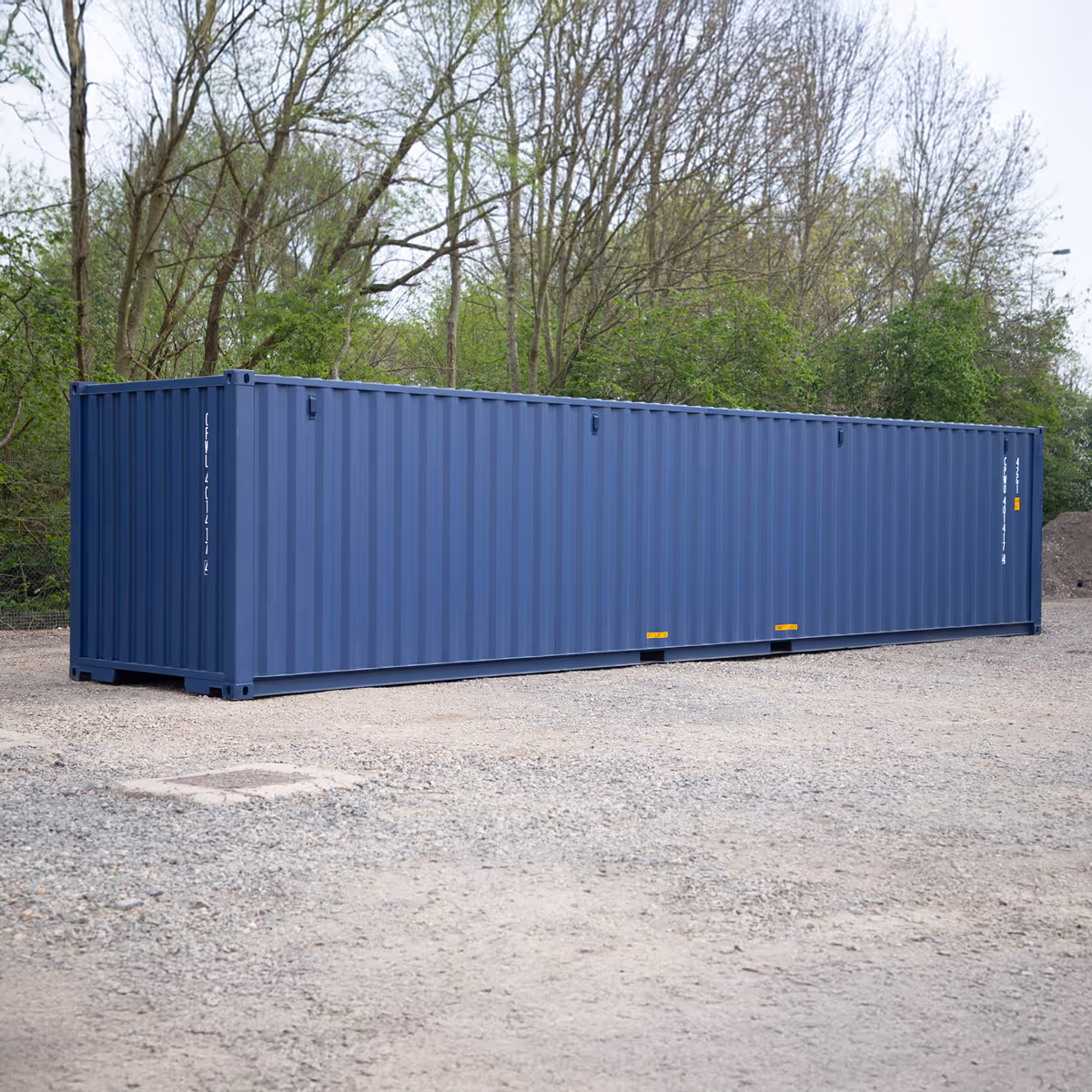 A large blue shipping container placed on a gravel surface with green trees in the background.