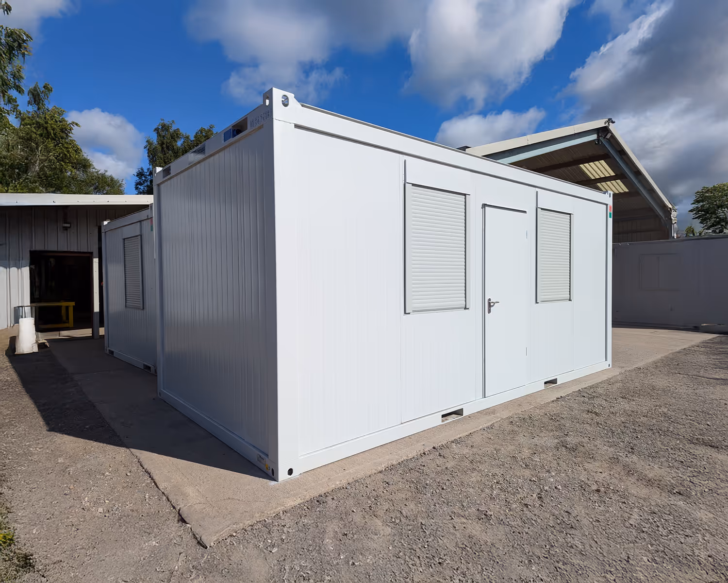 Two white prefabricated portable office units with closed windows and a door on a concrete and gravel surface under a partly cloudy sky.
