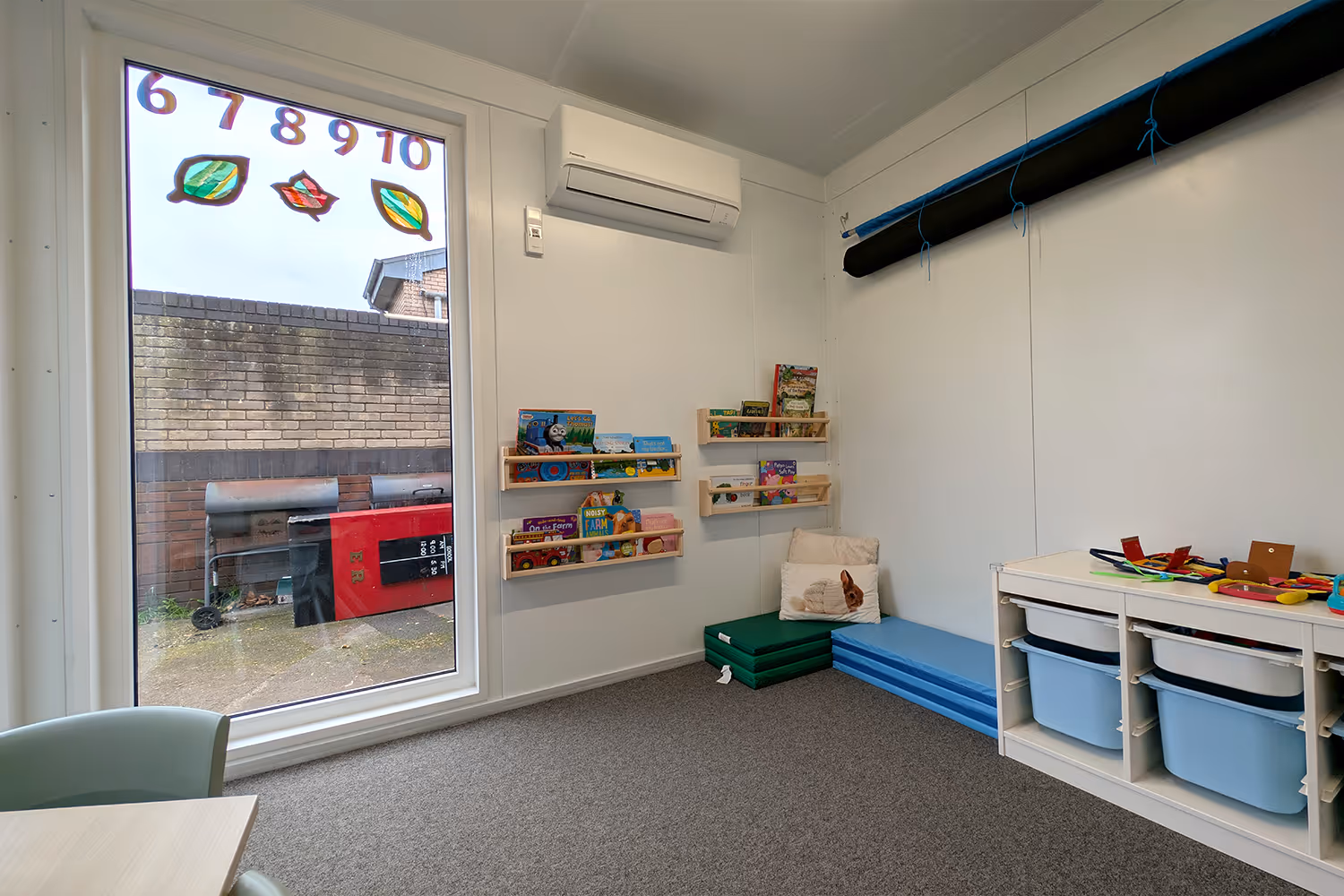 Bright classroom corner with carpeted floor, low shelves holding children's books and toys, mats with a cushion, and a large window with numbers 6 to 10 and leaf shapes on the glass.