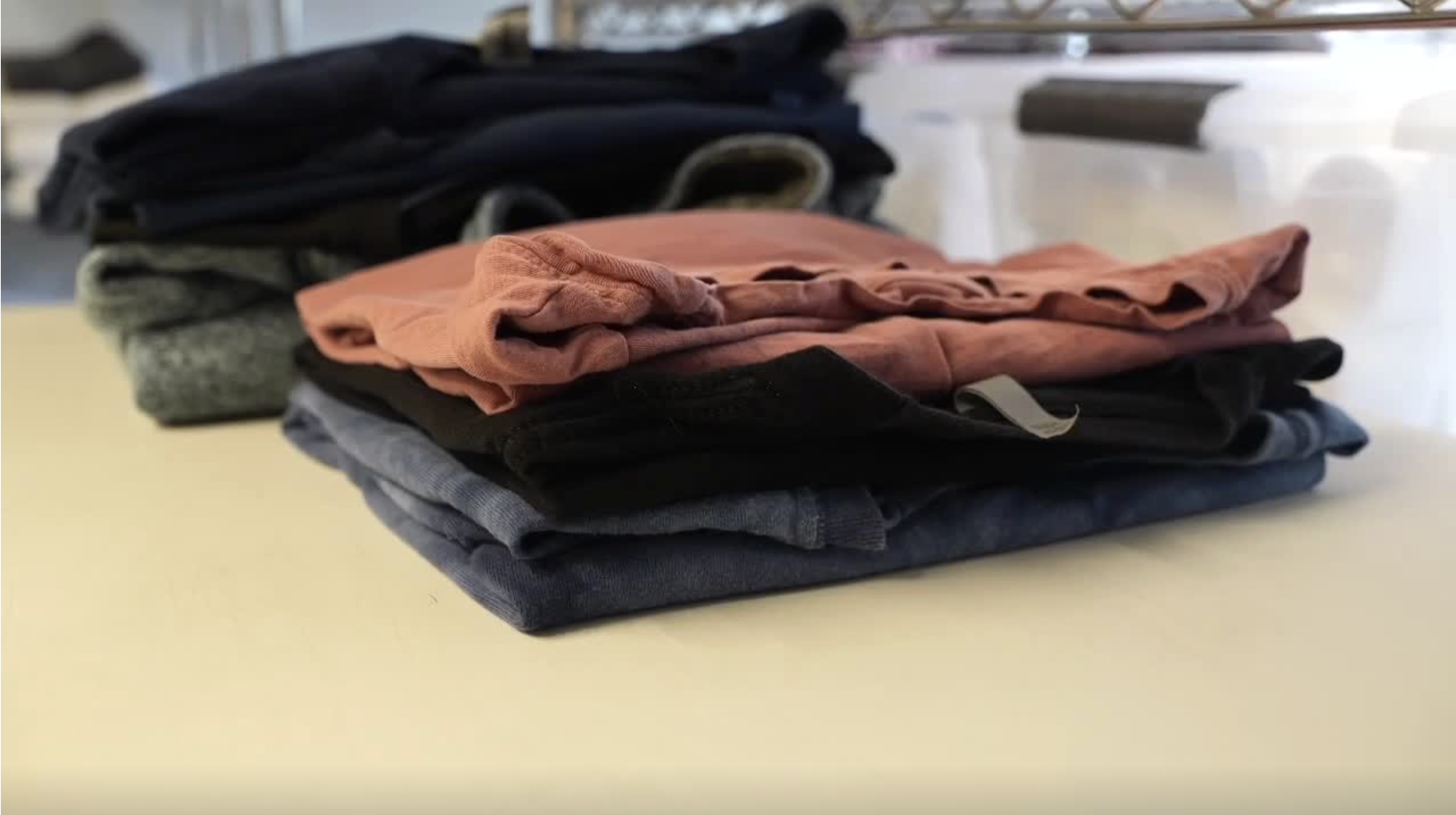 A neatly stacked pile of folded clothes in neutral and muted colors on a light-colored table, with storage boxes and shelves blurred in the background.