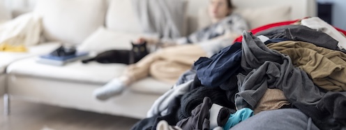 Pile of unfolded laundry in the foreground while a person rests in the background, illustrating how undone laundry lingers even when you're trying to take a break