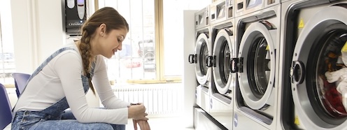 Person sitting alone in a laundromat at the end of a long day, suggesting the exhaustion of trying to fit laundry into a schedule that has no room for it
