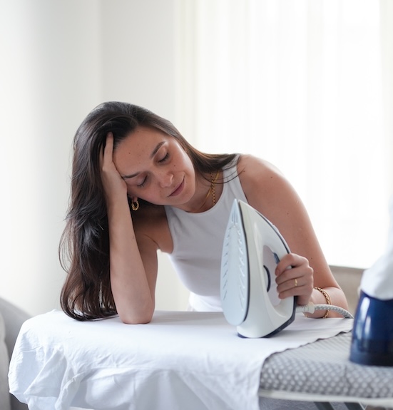 Woman standing at an ironing board with a distracted, weary expression, reflecting the mental weight of laundry that never stops demanding your time and attention