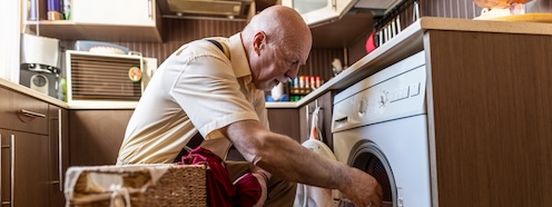 Older man bending down to reach a front-loading washing machine, illustrating the physical demands laundry places on seniors and the risk those demands quietly carry