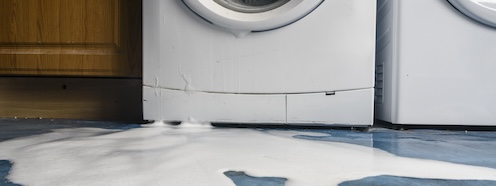 Water pooled on the floor in front of a washing machine, representing the sudden disruption of a broken appliance and the laundry problem it leaves behind