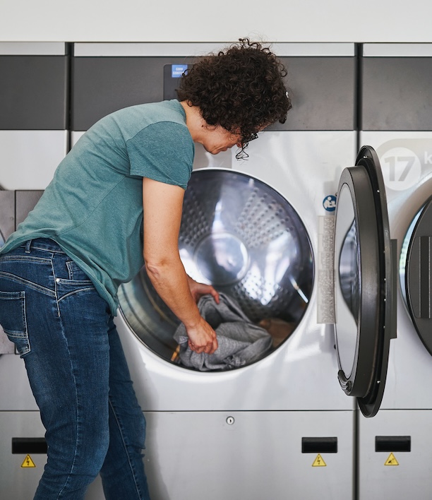 Person loading laundry into a machine at a laundromat, representing the recurring effort of handling laundry without in-unit machines at home