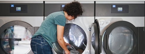 Person loading laundry into a machine at a laundromat, representing the recurring effort of handling laundry without in-unit machines at home