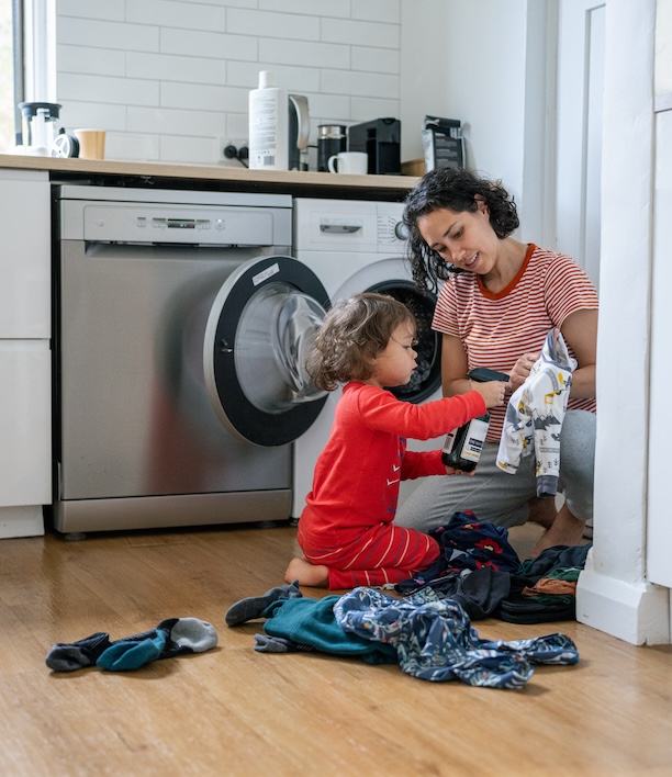 Parent showing a child an article of clothing while loading the washing machine together, representing how family laundry weaves into the constant demands of parenting