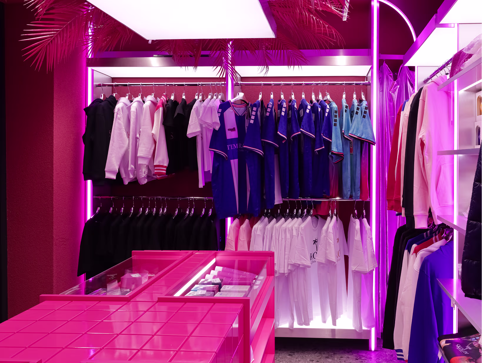 Strefa odzieżowa butiku Stacja Skolim z wieszakami koszulek i bluzy, różowe neonowe słupy i palmy