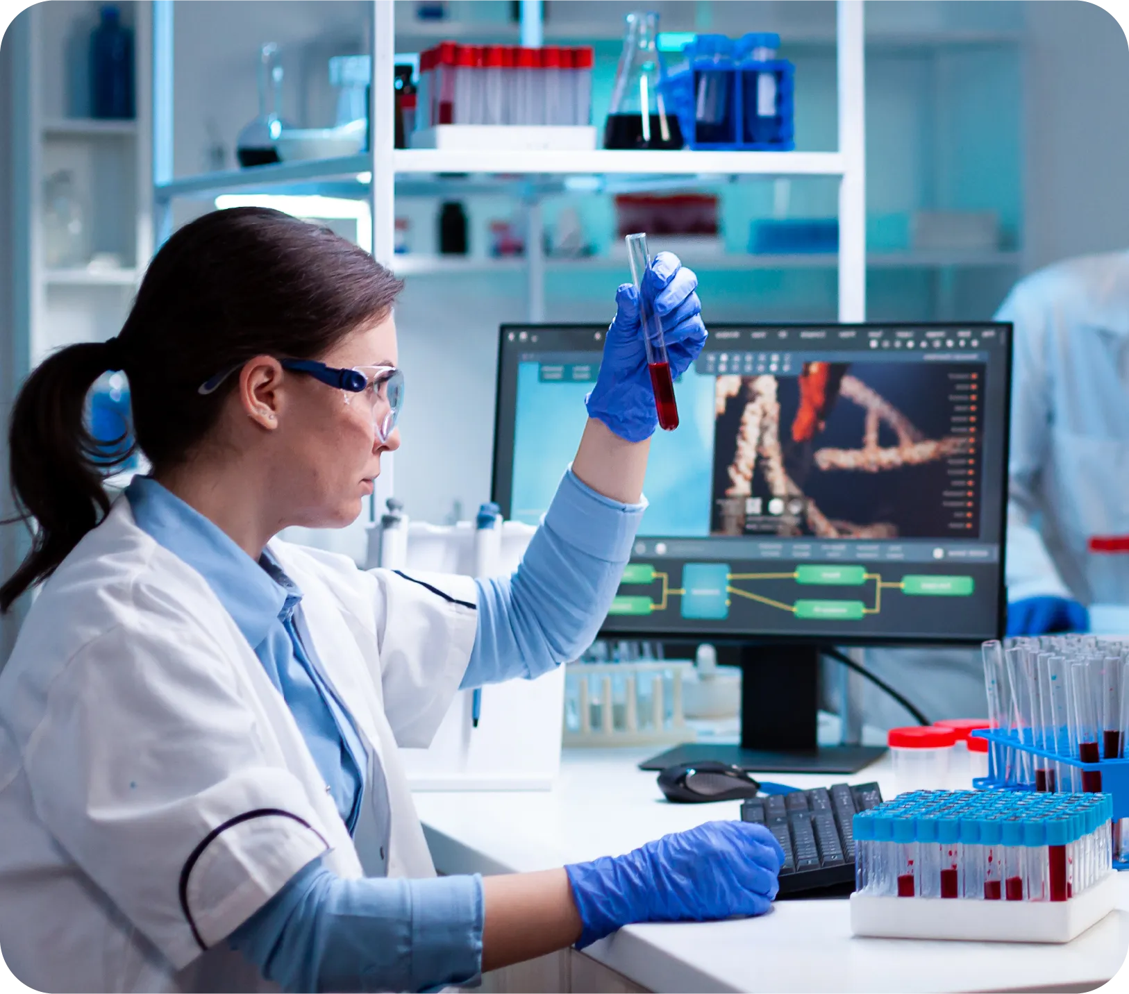 Female scientist wearing protective glasses and gloves examining a test tube with red liquid in a laboratory with DNA imagery on a computer screen.