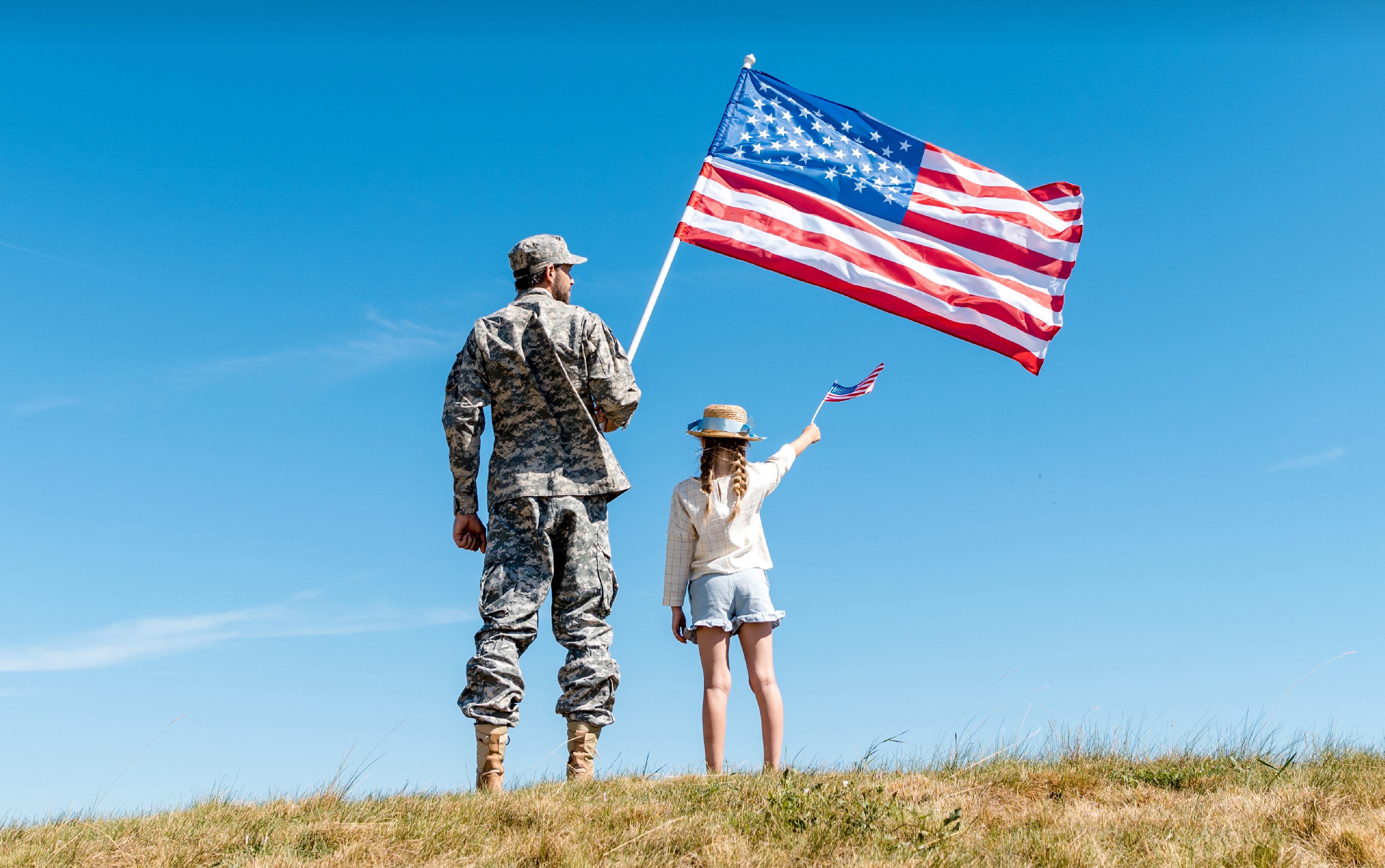 Soldier and young girl standing on hill holding large and small American flags under blue sky.