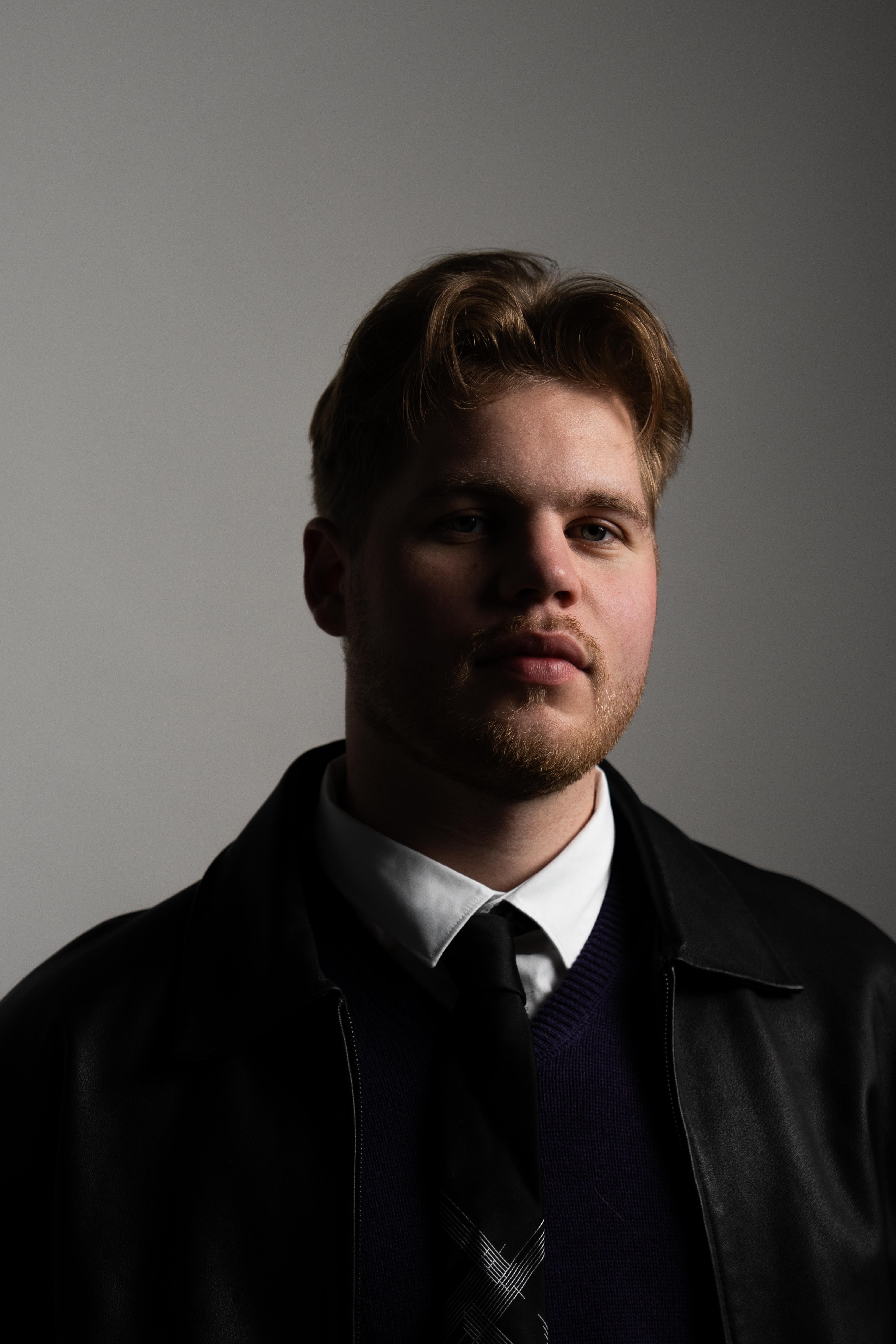 Young man with light brown hair and beard wearing a white shirt, black tie, navy sweater, and black jacket against a gray background.