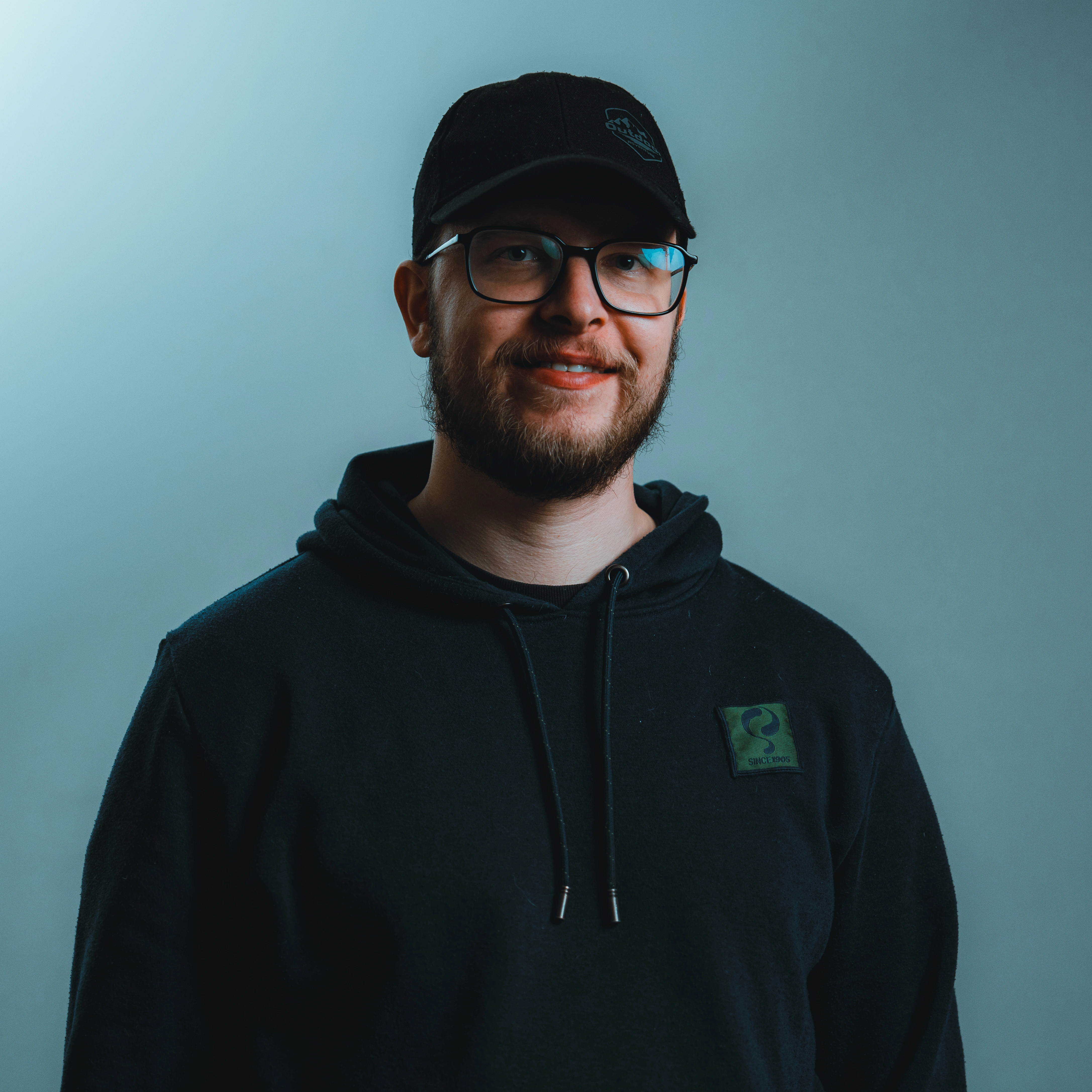 Man with a beard wearing glasses, a black baseball cap, and a black hoodie smiling against a plain blue-gray background.