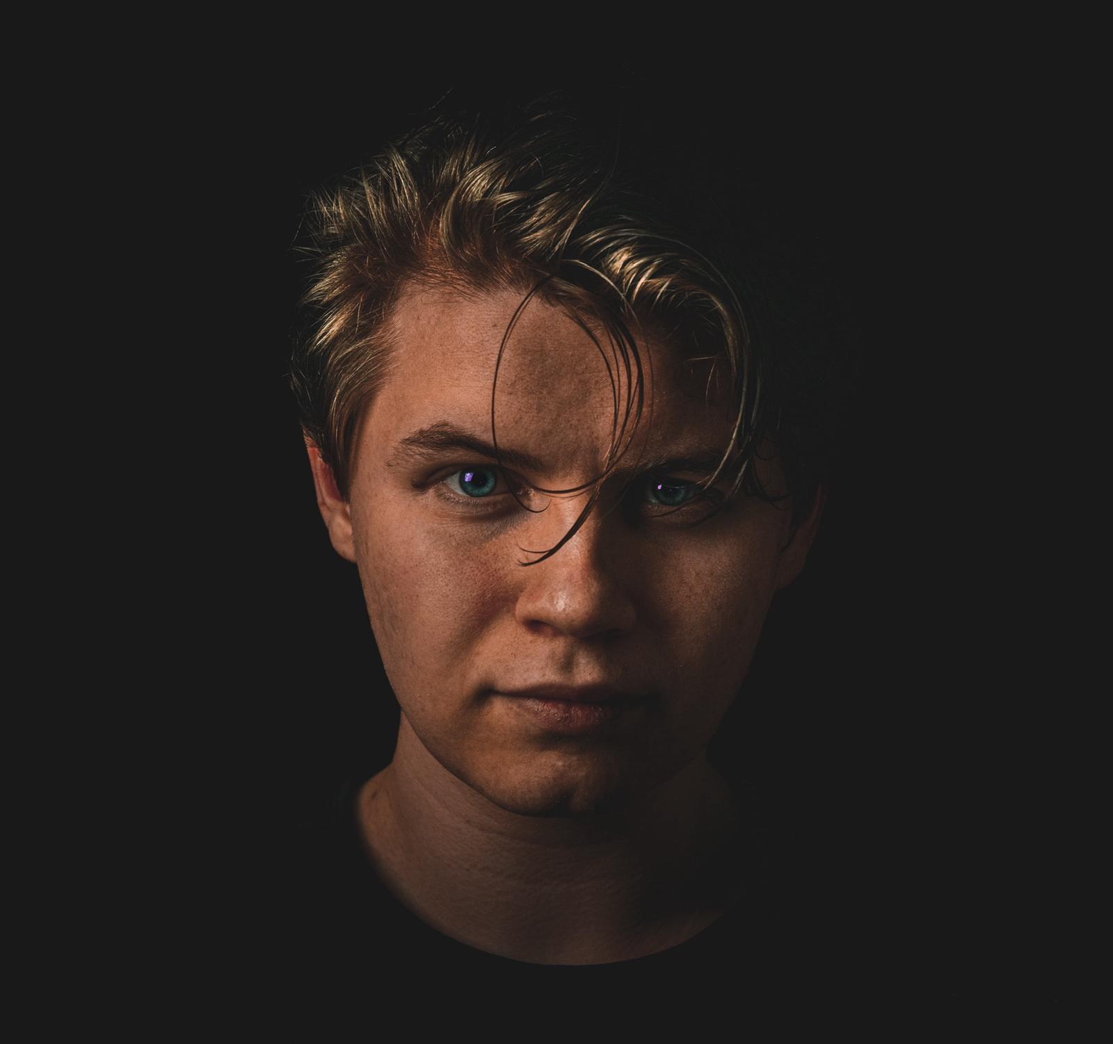 Close-up portrait of a young man with light brown hair and green eyes against a black background.