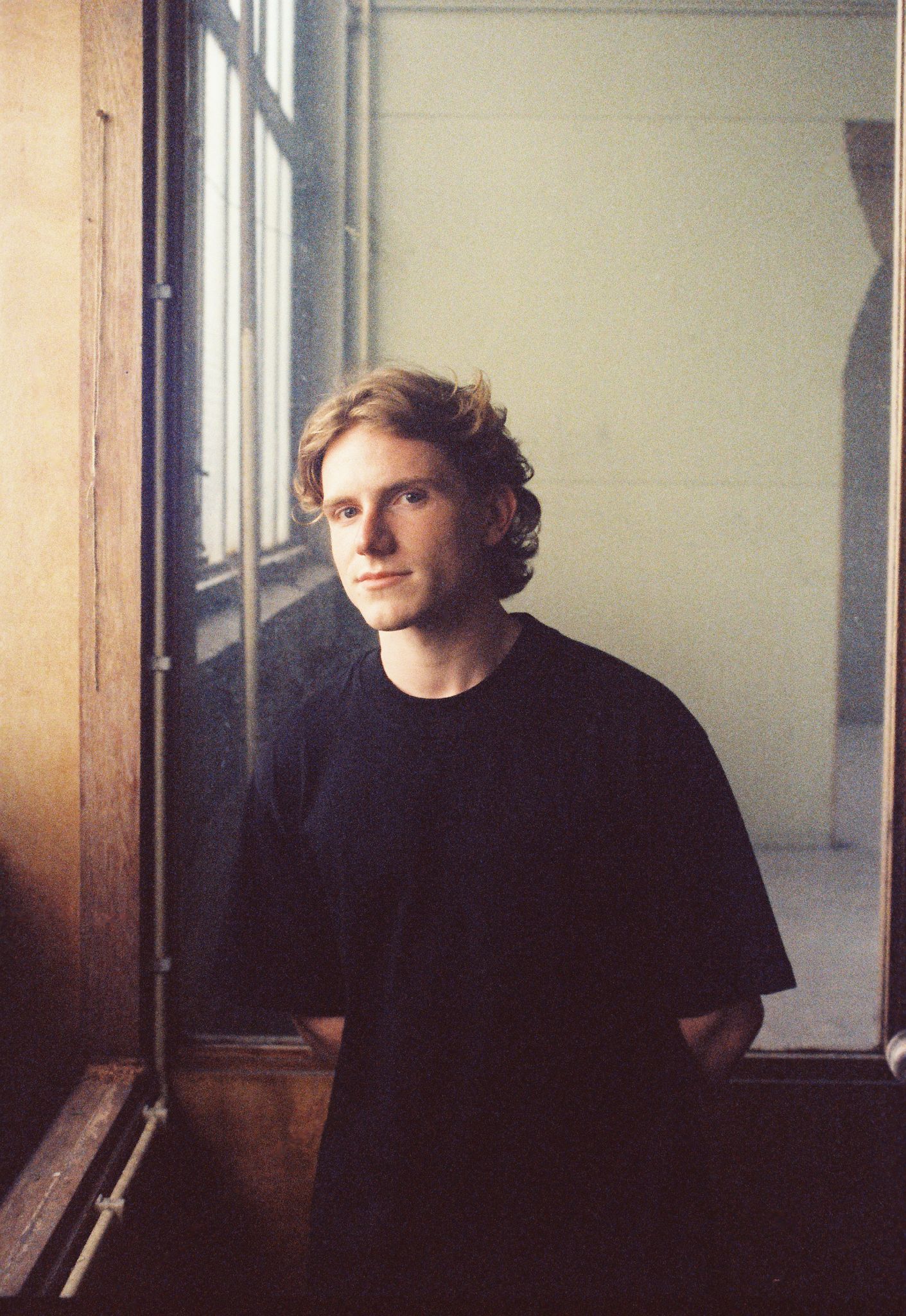 Young man with curly blonde hair wearing a black t-shirt standing indoors near a window with natural light.