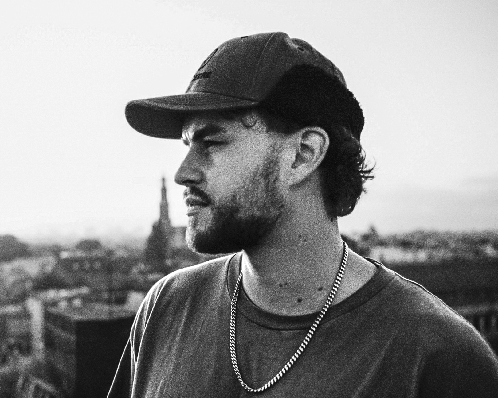 Black and white side portrait of a bearded man wearing a cap and chain necklace, looking into the distance against a blurred cityscape background.