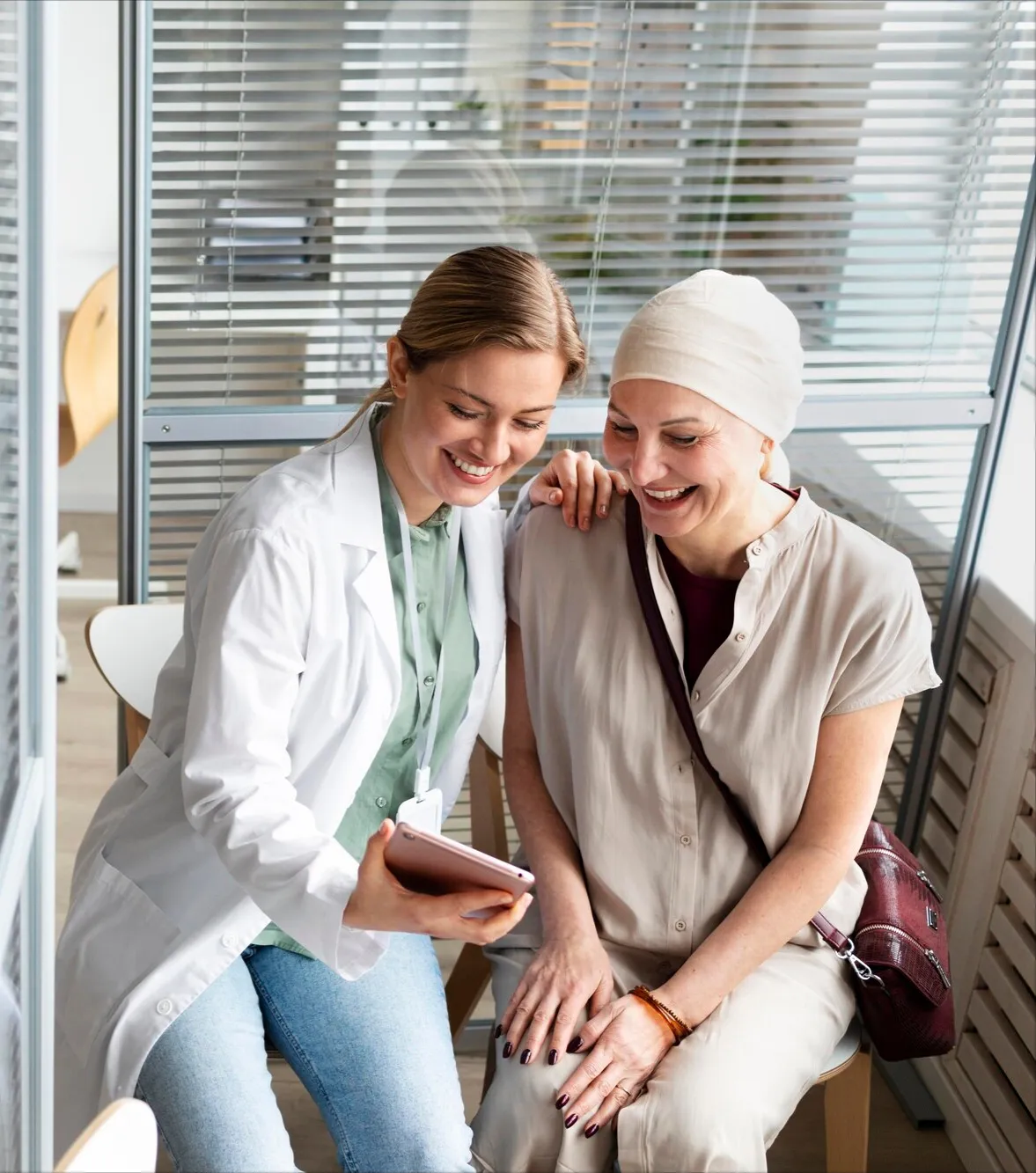 A female physician shares information on a rose-gold tablet. The patient, wearing a light headscarf, is looking on and smiling. They are sitting in an office setting.