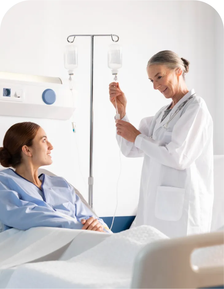A medical professional adjusts an IV line while a patient in a hospital gown looks on.