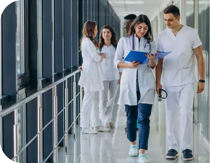 Healthcare professionals are in the hospital hallway. A doctor and nurse are reviewing a blue patient chart as they walk.
