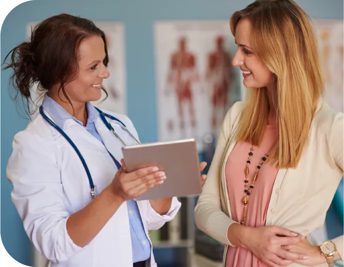 A female doctor wearing a white coat and stethoscope holds a tablet while speaking with a smiling patient in a medical office. She is wearing a pink blouse and a white sweater.