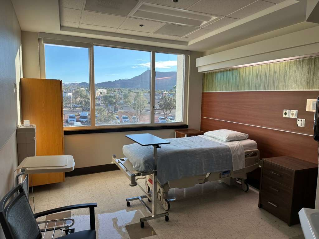A hospital room with a single bed neatly made, positioned by a large window showing a scenic mountain view. The room feels calm and orderly.