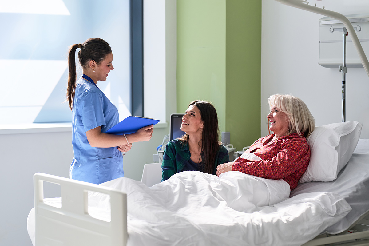 A nurse in blue scrubs holds a clipboard, talking to a smiling woman beside an older woman in a hospital bed. The scene conveys warmth and care.