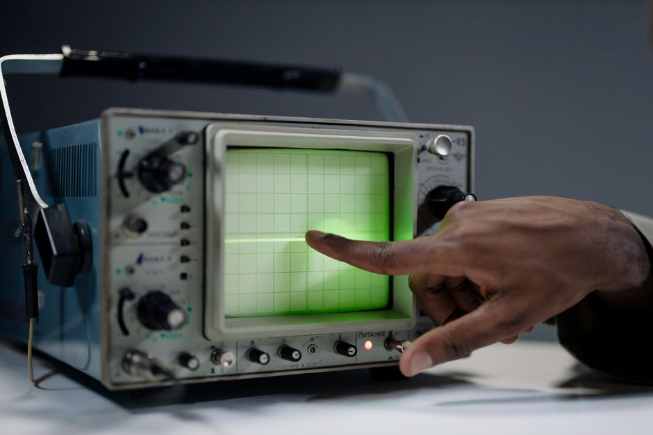 A hand pointing at the screen of a vintage oscilloscope indoors.