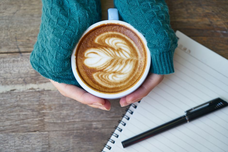 Cozy latte art in hands wearing knitted sweater, notebook and pen on wooden table.