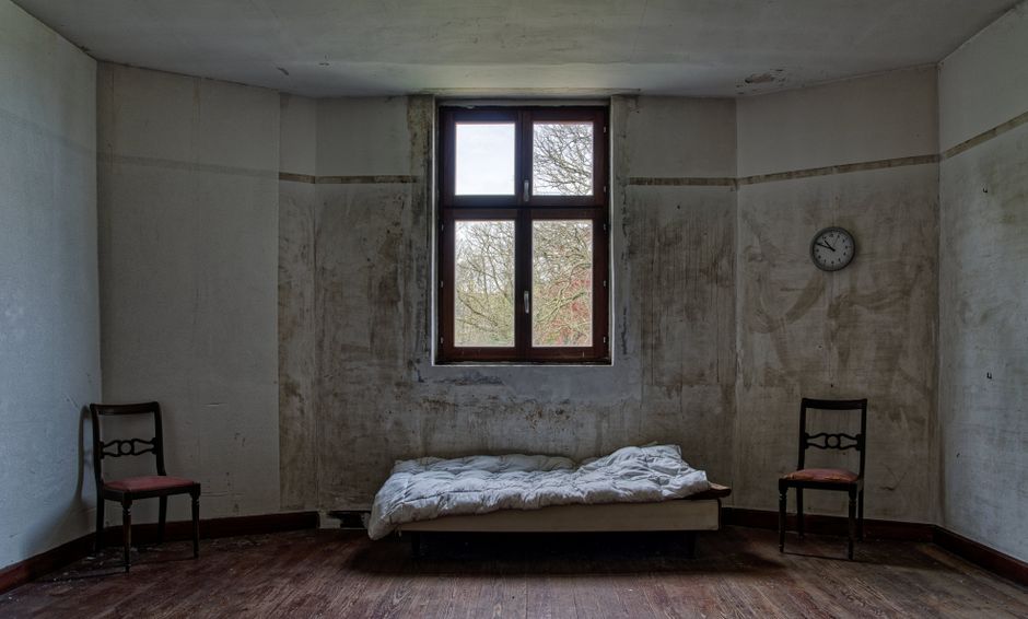 An old, abandoned room with a single bed, two chairs, and a window.