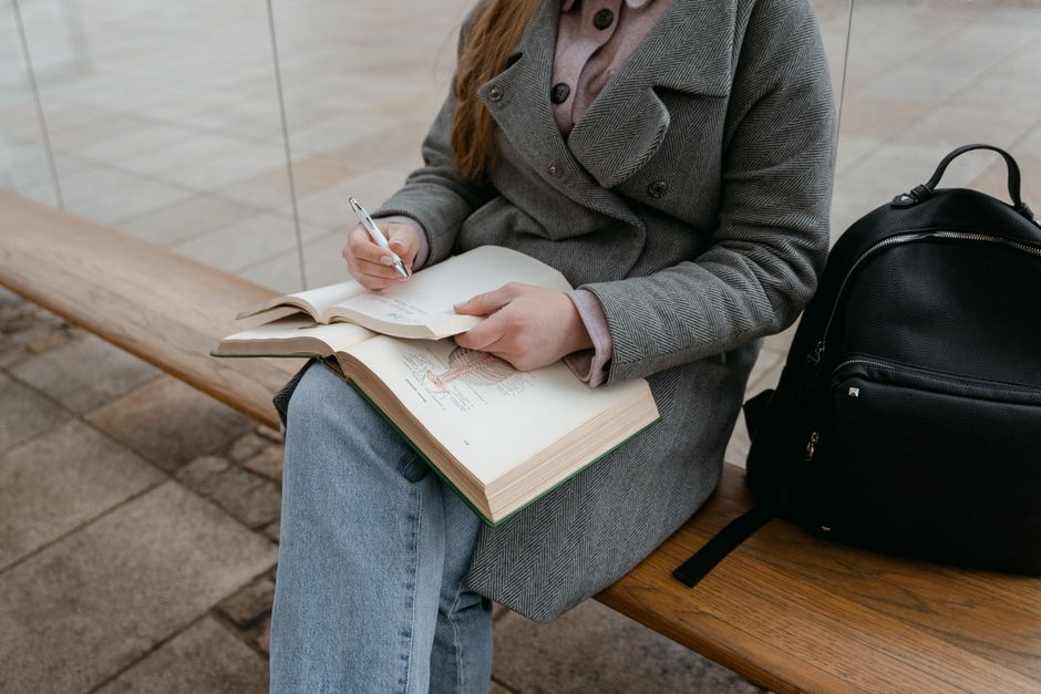 A woman in a gray coat sitting on a bench writing in a notebook outdoors.