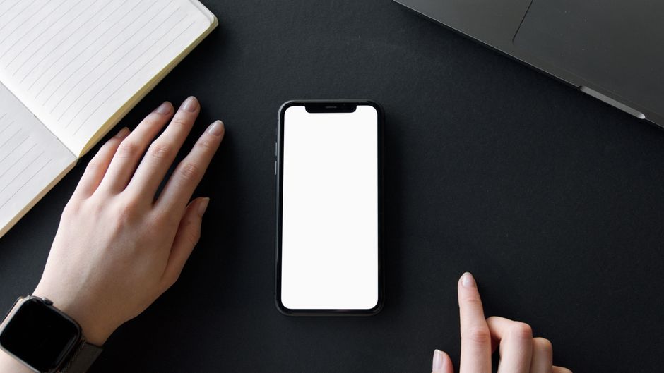 A minimalist workspace featuring a smartphone mockup, hands, and a notebook on a black surface.