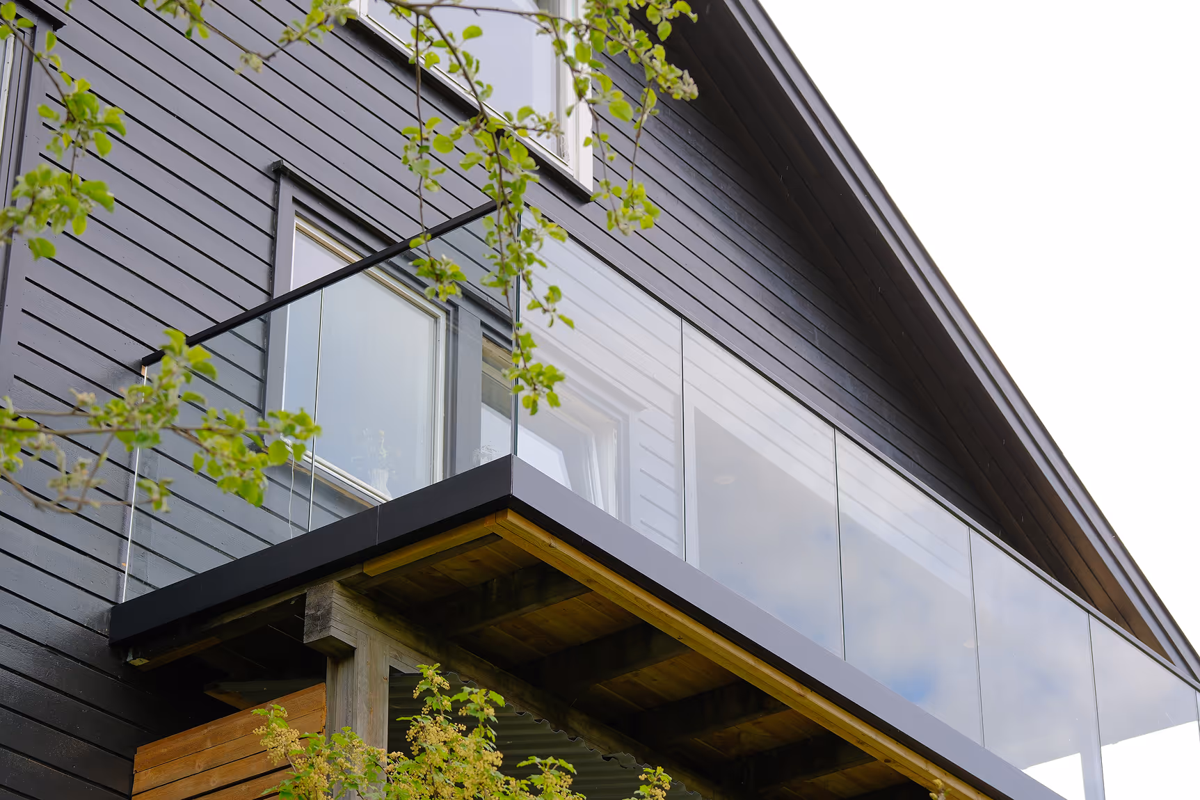 Modern house balcony with glass railing and black wooden siding, partially framed by green tree branches.