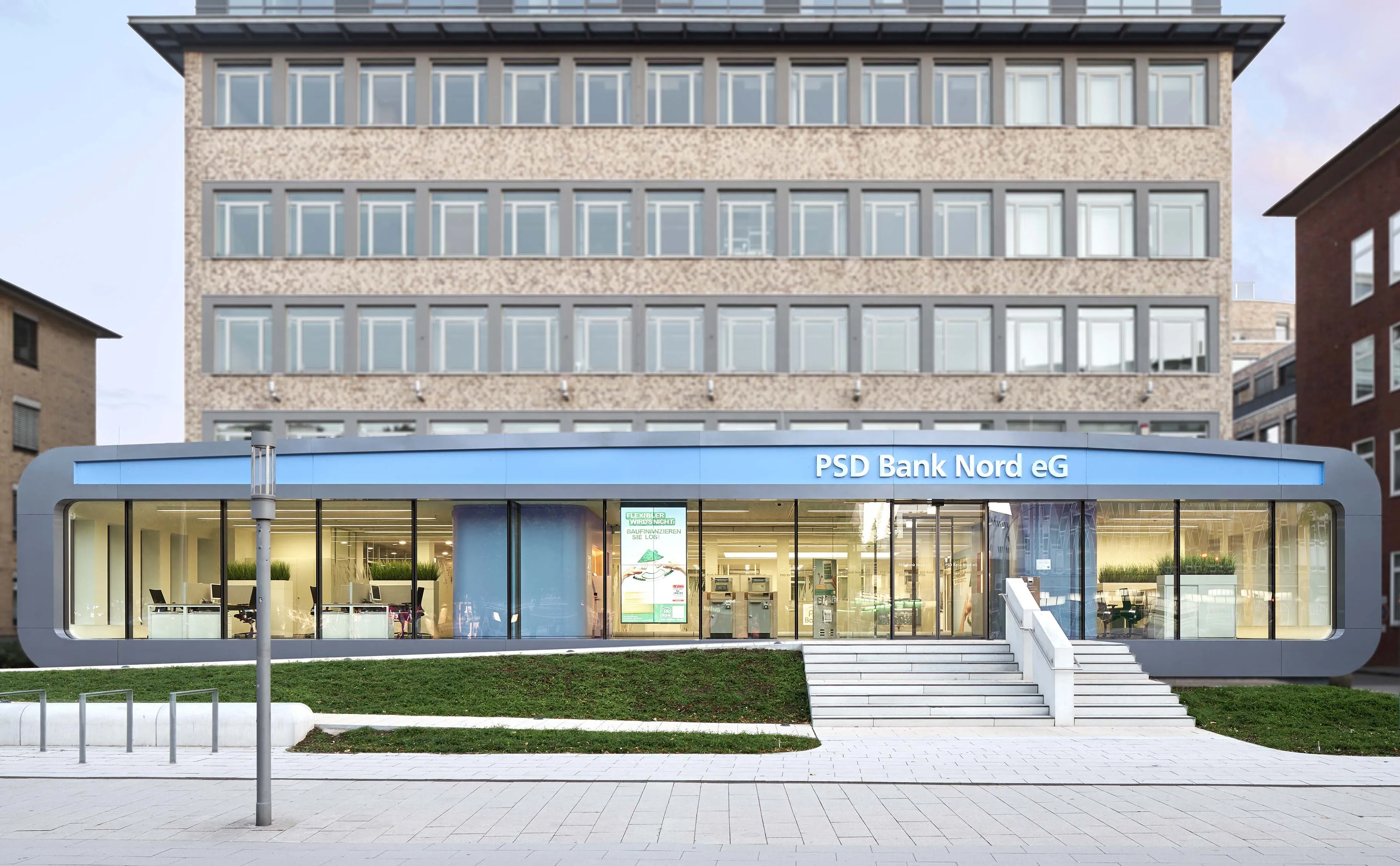 Moderner Eingang der PSD Bank Nord eG mit beleuchteten Glasfenstern und Treppe vor einem mehrstöckigen Backsteingebäude.