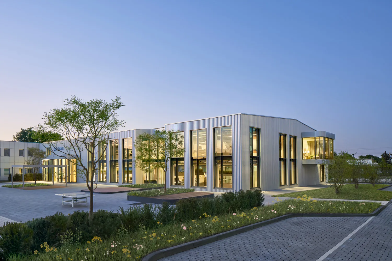 Modernes Bürogebäude mit großen Fenstern, Bäumen, Blumen und gepflastertem Parkplatz bei Sonnenuntergang.